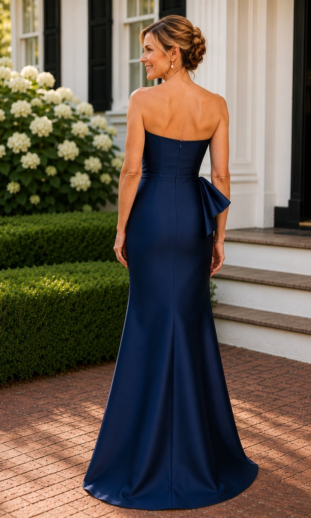 A woman stands outdoors in a navy blue Strapless Bow Mermaid Formal Dress: Rina Di Montella R113, a strapless mermaid gown with a floor-length hem and bow detail at the back.